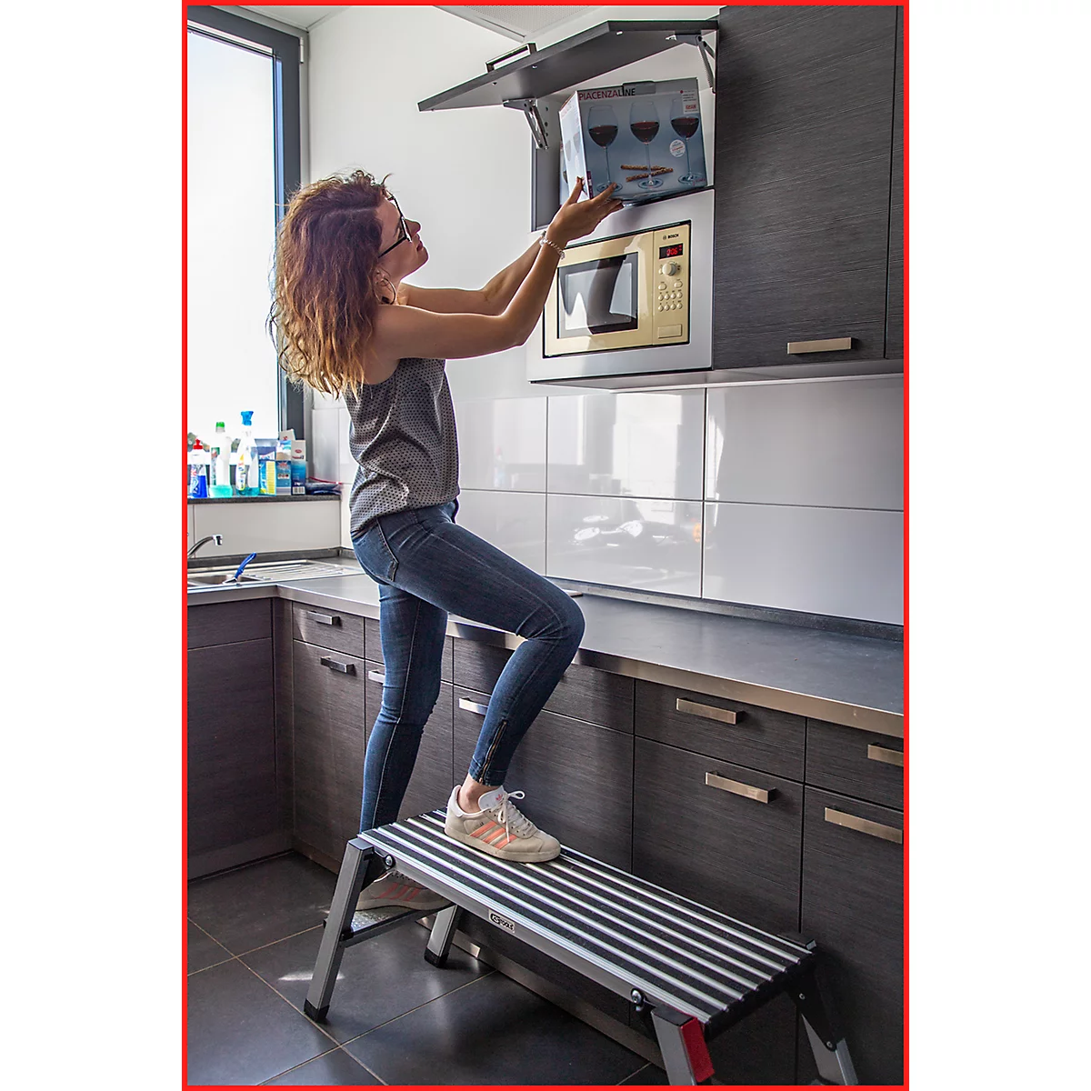 Frau in Jeans und Top steht auf Tritt, um Schrank über Mikrowelle zu öffnen. Küche im Hintergrund.